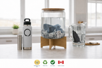 Glass water filter with rocks on a wooden stand, surrounded by water bottles and a pitcher on a light-colored surface.